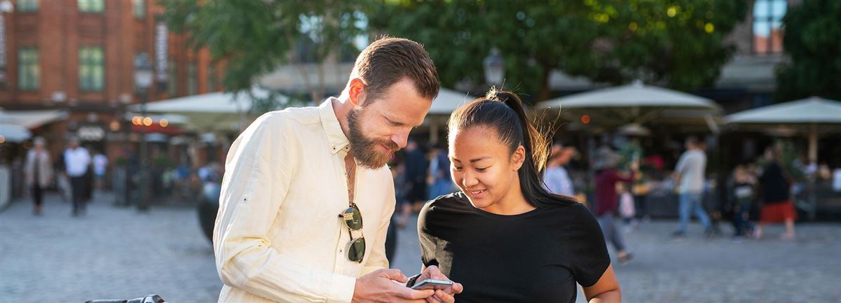 En man och en kvinna står utomhus i stadsmiljö och tittar på något i mannens mobiltelefon.