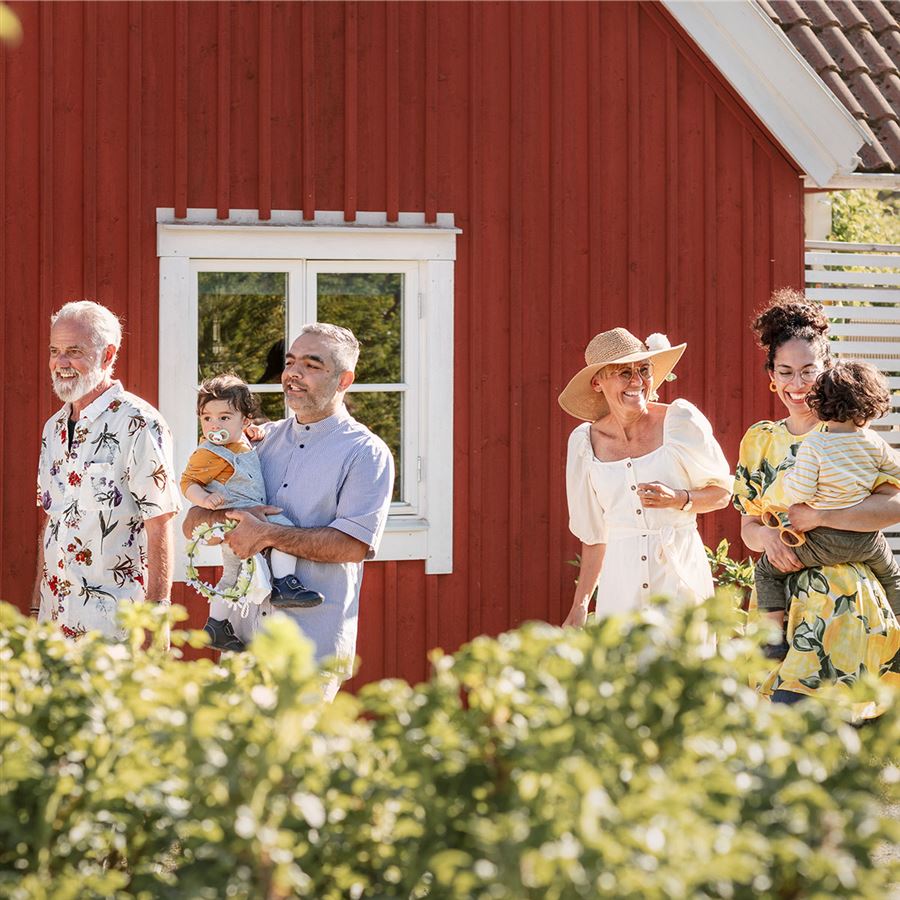 En familj står framför en rödmålad stuga i strålande sol.