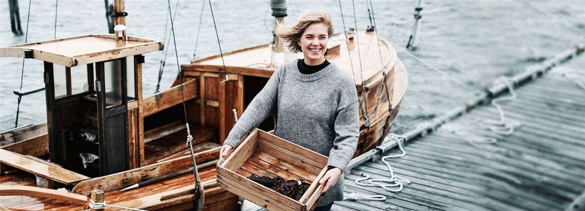 Kvinna med låda i händerna framför en fiskebåt funderar på sin tjänstepension