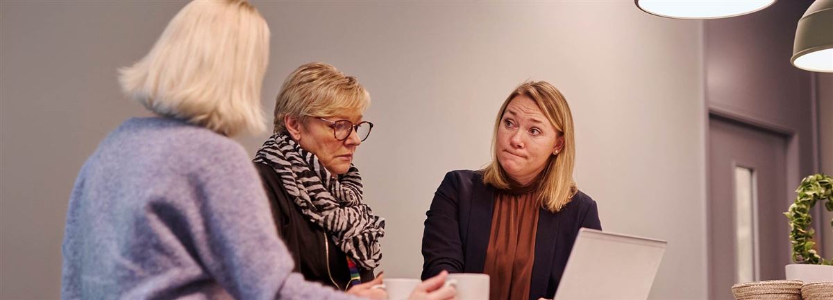 Tre kvinnor står vid en dator på en arbetsplats och diskuterar något de ser på skärmen.