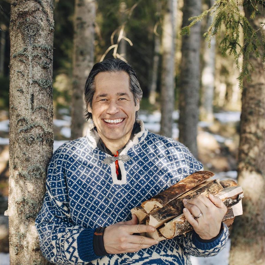 Medelålders man lutar sig mot en trädstam i skogen. Han håller ved i famnen och ler mot kameran.