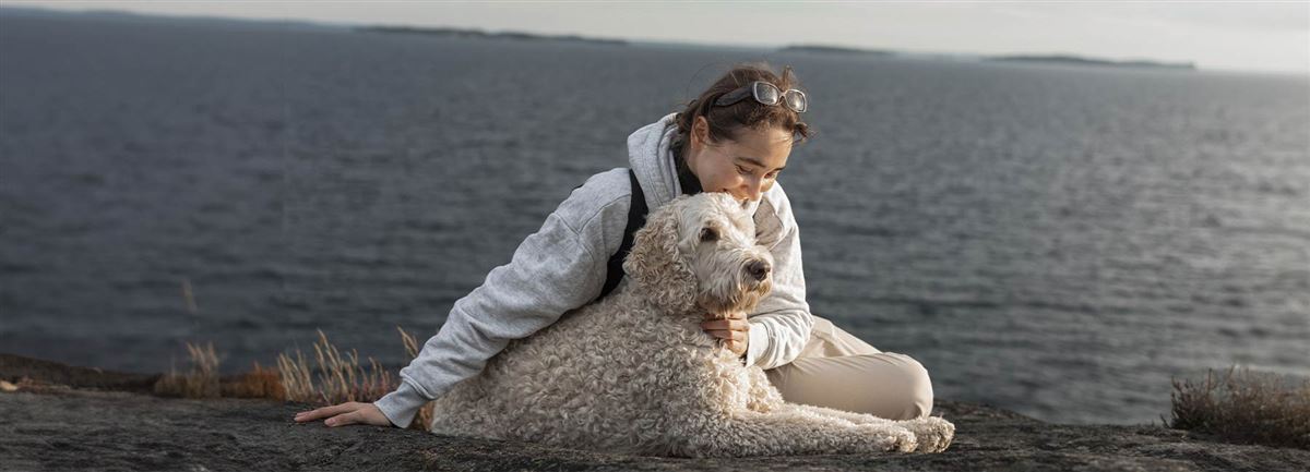 Kvinna sitter på en klipphäll vid havet. Hon har en stor vit hund bredvid sig som hon klappar.