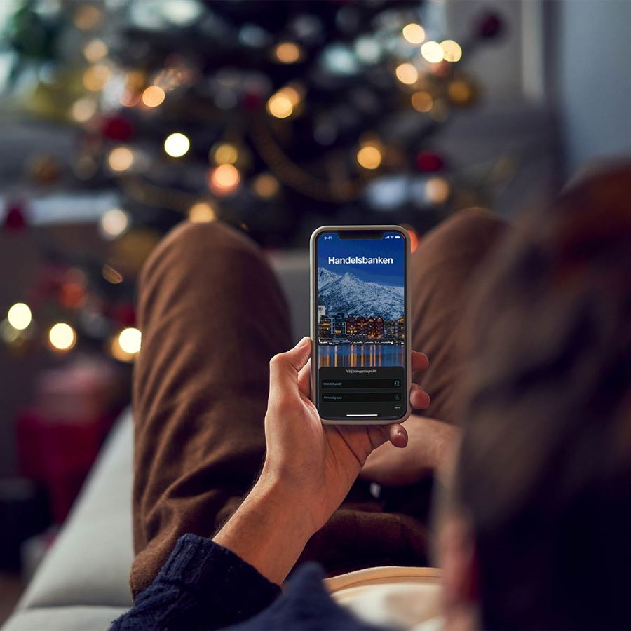Person som ligger i soffan och har Handelsbankens företagsapp på sin mobil. En julgran i bakgrunden.