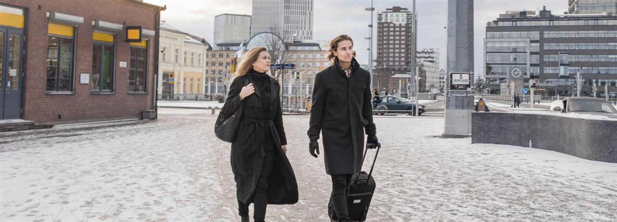 Kvinna och man på affärsresa går över ett torg i en stad. Mannen drar en kabinväska. 