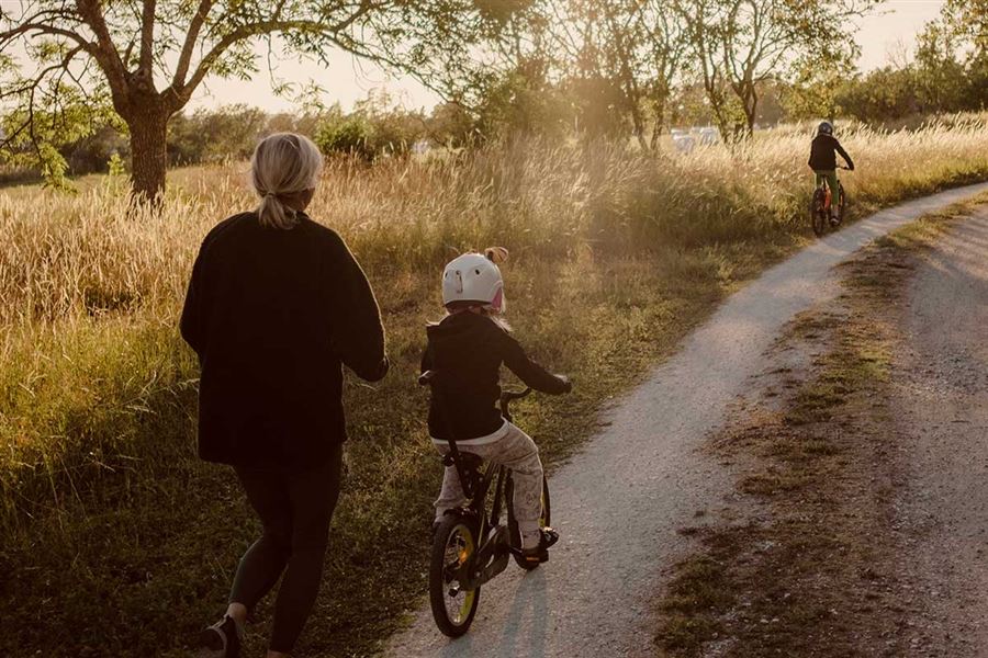 En kvinna springer på en grusväg bredvid ett fält. Två barn cyklar framför henne. 