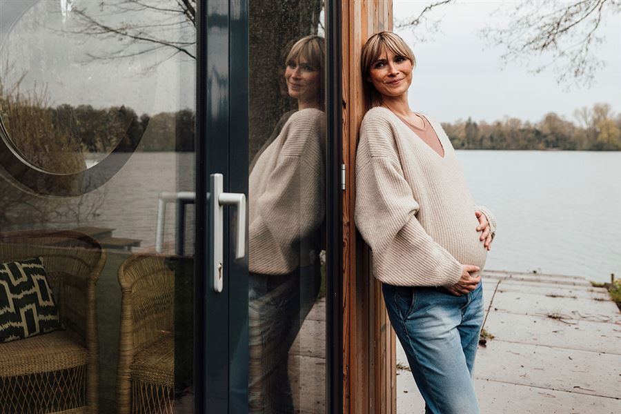 Leende höggravid kvinna lutar sig ledigt mot husväggen.