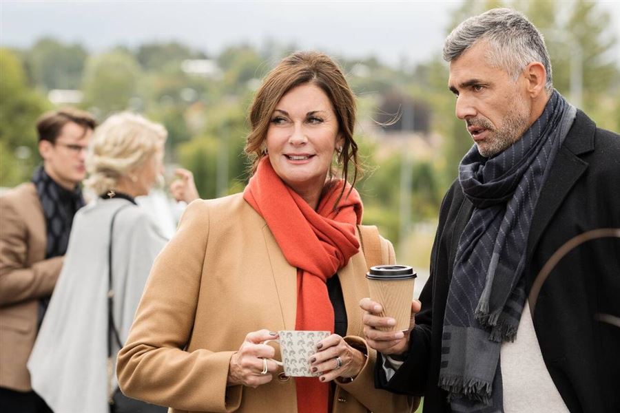 En kvinna och man står utomhus och pratar. Kvinnan har en kopp kaffe i handen.