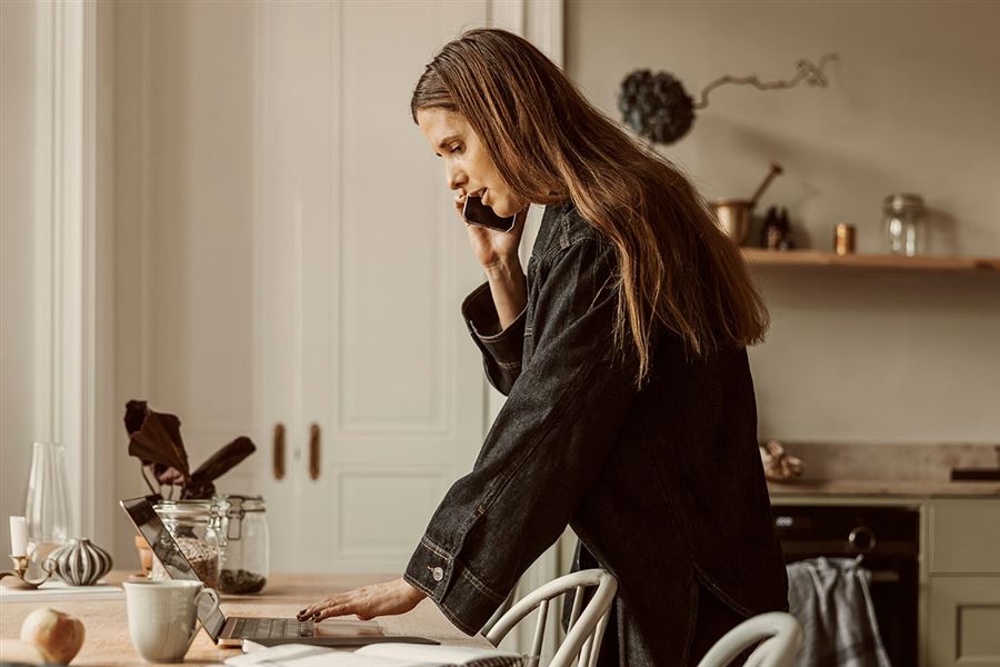 En kvinna står upp och tittar på sin laptop medan hon pratar i mobiltelefon.