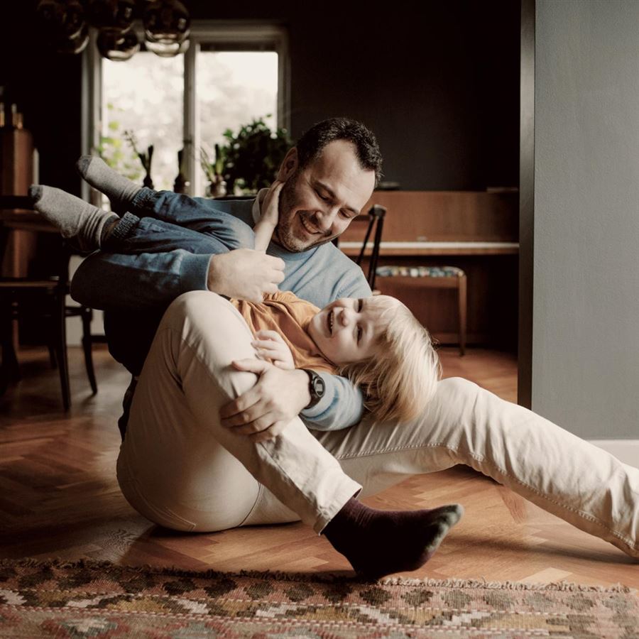 Förälder som sitter på golvet och leker med ett skrattande barn i vardagsrummet.