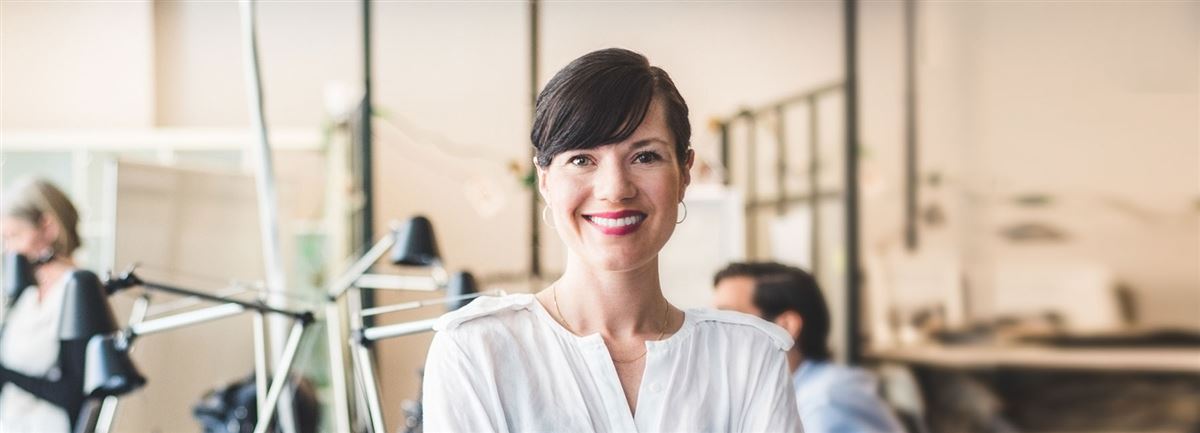 Business woman is in the office, she is smiling. In the background people are working. 