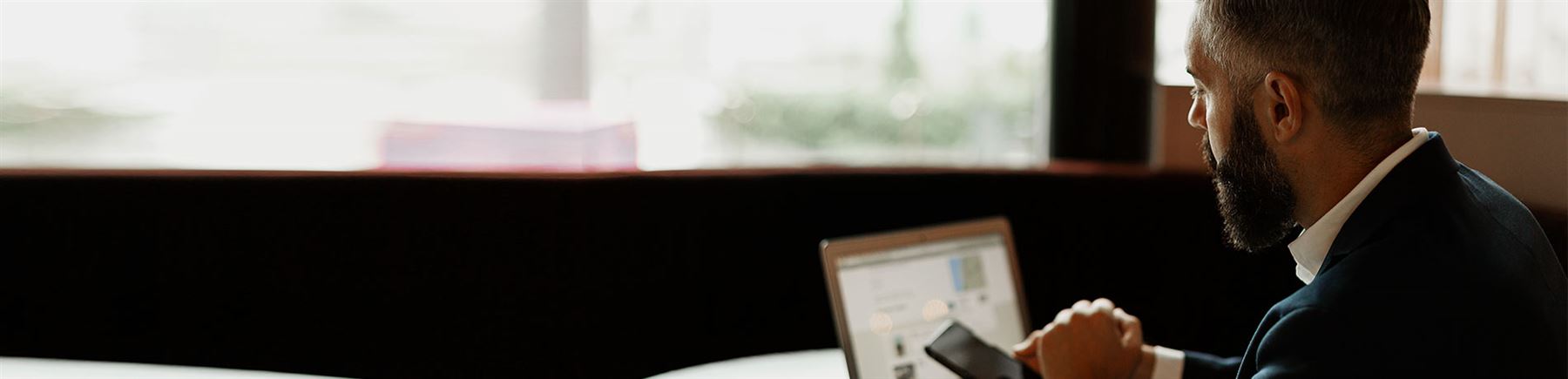 Man sits by his laptop with a coffee. He has a mobile phone in his hand.