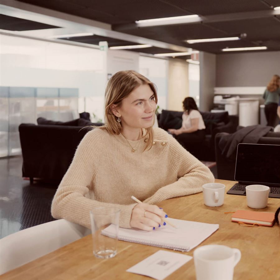 A woman is sitting at a meeting in an office. She is smiling and taking notes. 